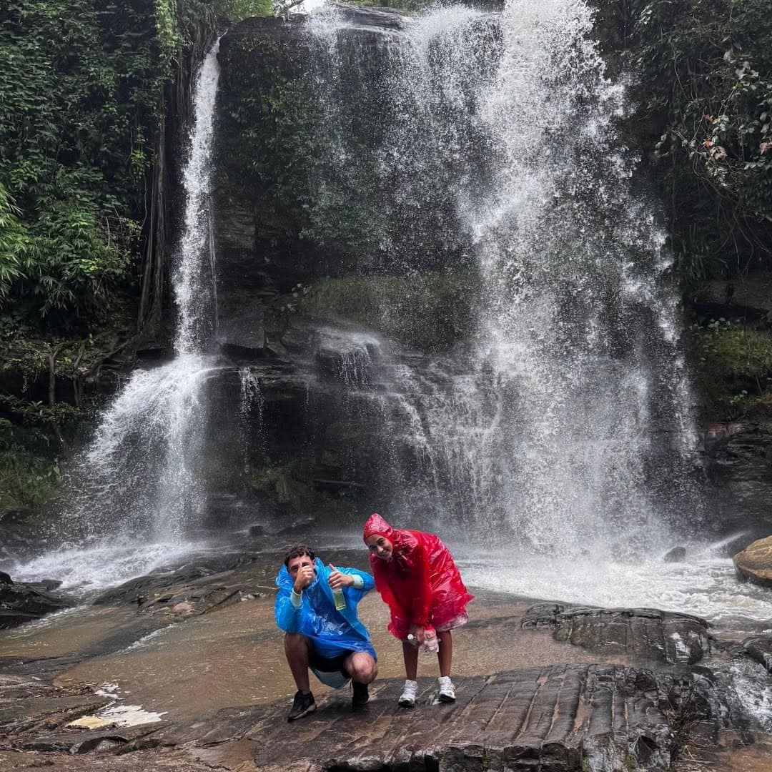 Two guests in raincoats stand on wet rock at the base of a forest waterfall